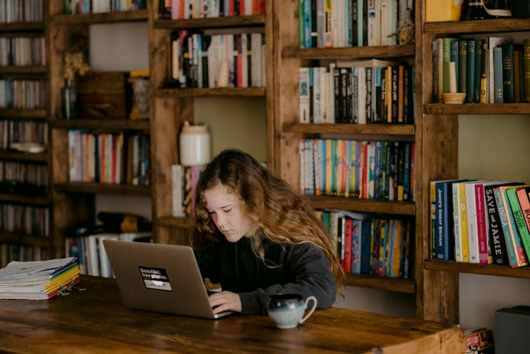 A young girl works at a laptop, with bookcases behind her, lined with books.