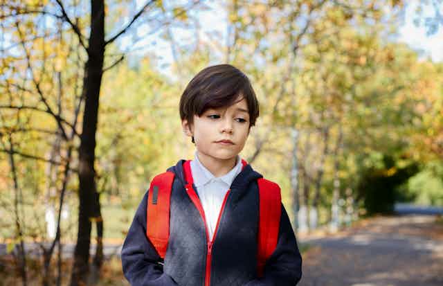 A young boy in school jumper and backpack, walks on a road with trees in background. He has a downcast expression.