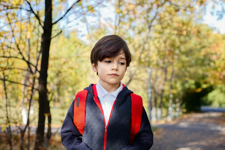 A young boy in school jumper and backpack, walks on a road with trees in background. He has a downcast expression.