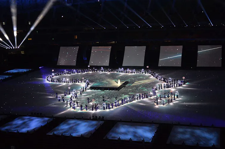 A sport stadium with huge screens showing the cosmos. On the field people hold lights and form the shape of the African continent.