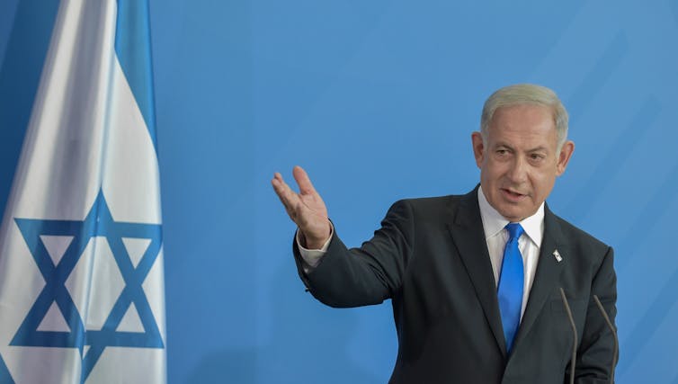Benjamin Netanyahu speaking in front of an Israel flag with his right hand outstretched.