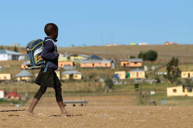 A barefoot child wearing a dark blue uniform, a rucksack on their back, walks past a group of simple houses