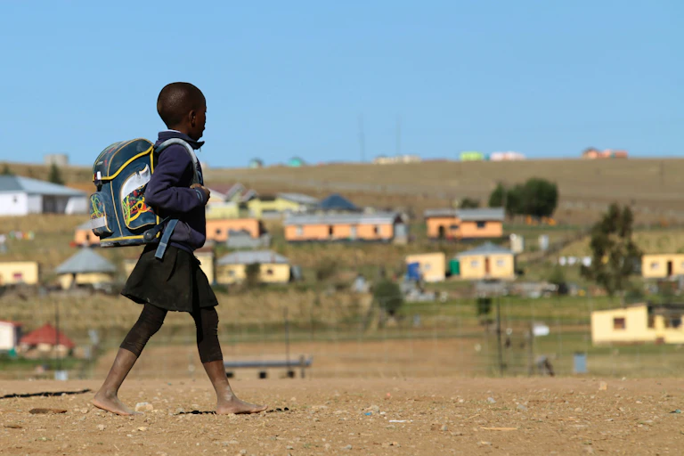 A barefoot child wearing a dark blue uniform, a rucksack on their back, walks past a group of simple houses