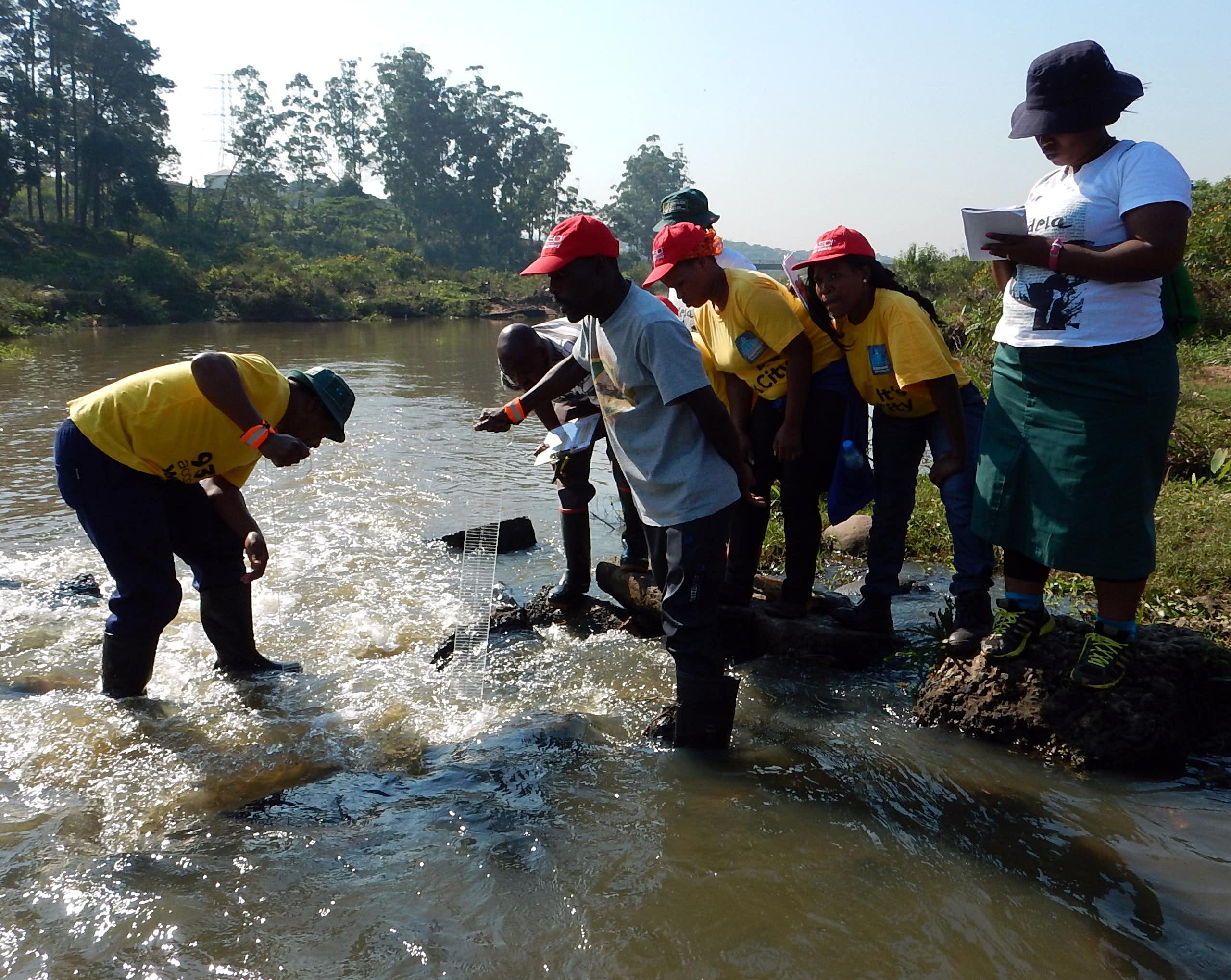 A group of young people in yellow tshirts and red caps stand in a shallow stream using equipment to check the water quality