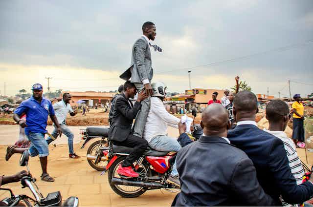 A man in a suit stands on the back of as moving motorbike, held from behind by another man as they drive along a dirt road in Uganda, some people running alongside or watching on.