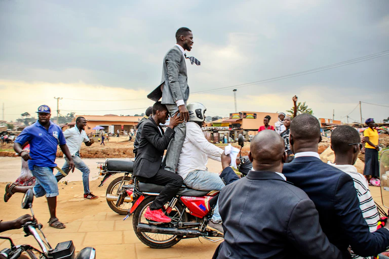 A man in a suit stands on the back of as moving motorbike, held from behind by another man as they drive along a dirt road in Uganda, some people running alongside or watching on.