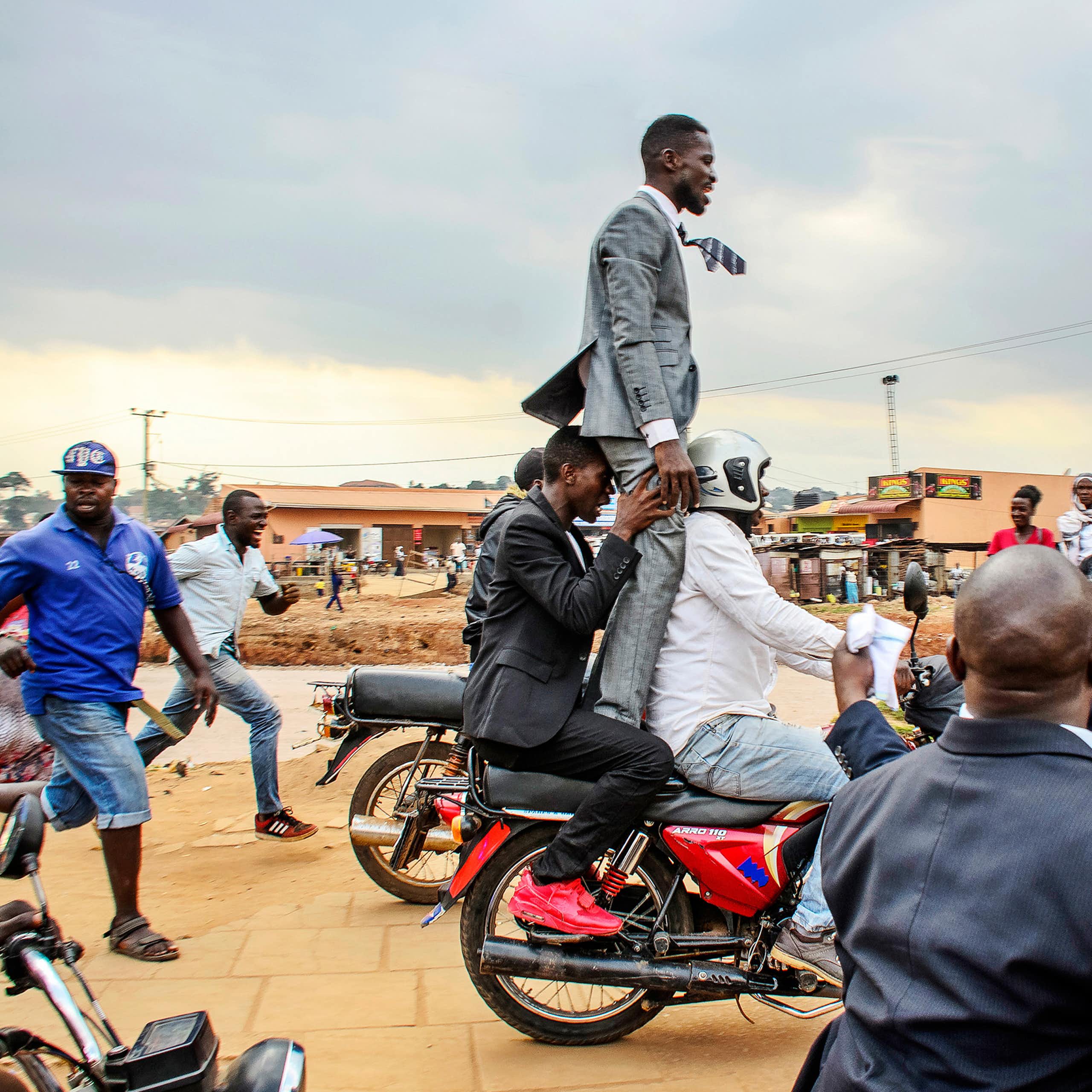 A man in a suit stands on the back of as moving motorbike, held from behind by another man as they drive along a dirt road in Uganda, some people running alongside or watching on.