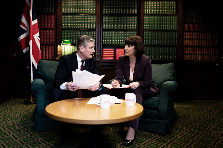 Keir Starmer and Rachel Reeves sitting on a sofa, looking at each other, with papers spread across the table in front of them.