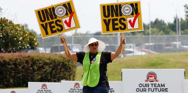 A woman holds placards with the words 'Union Yes' aloft in both hands while walking by yard signs saying 'vote no August 3-4.'