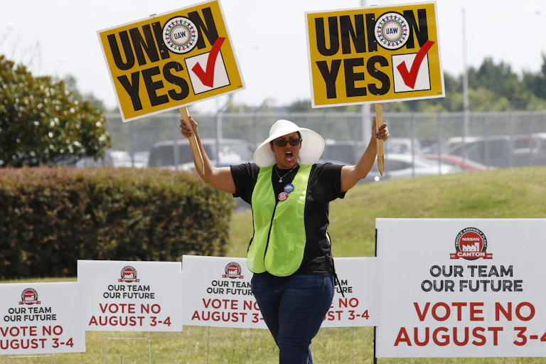 A woman holds placards with the words 'Union Yes' aloft in both hands while walking by yard signs saying 'vote no August 3-4.'