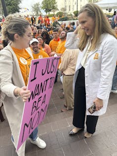 A woman in a long white sweater, holding a pink sign that says 'I just want to be a mom,' speaks with another blonde woman in a doctor's coat.