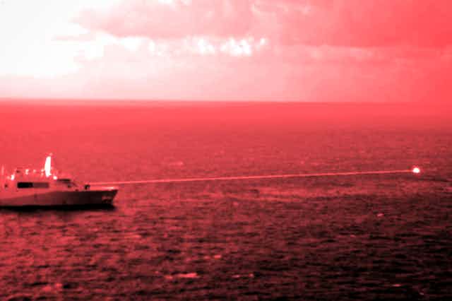 a red-tinted photo of a warship at sea firing a laser beam
