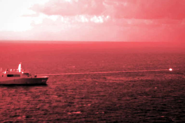 a red-tinted photo of a warship at sea firing a laser beam