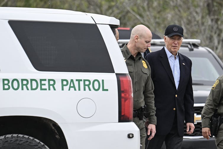 Joe Biden wears a black blazer and a black hat as he stands next to a bald white man wearing a green uniform and a white truck that says 'Border Patrol' in green