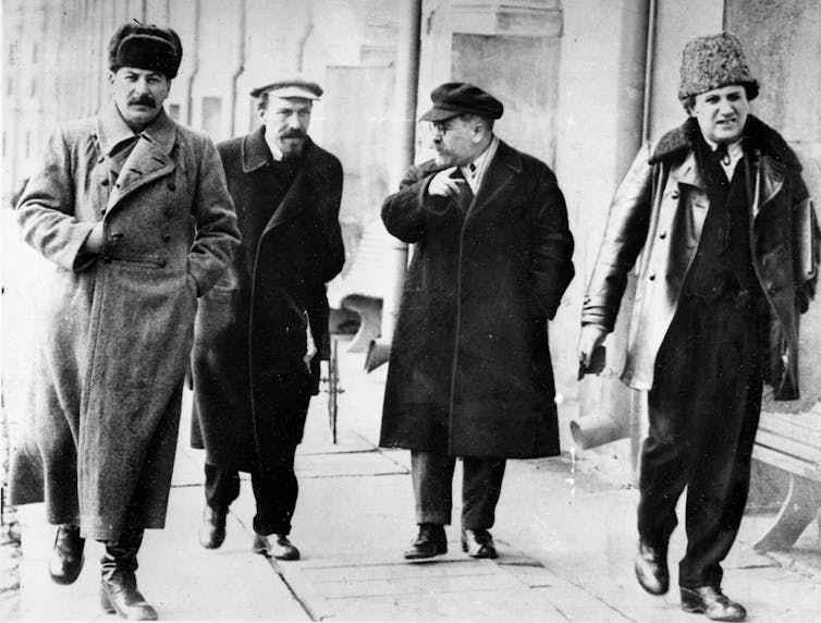 Four men in hats and overcoats walk down a street.
