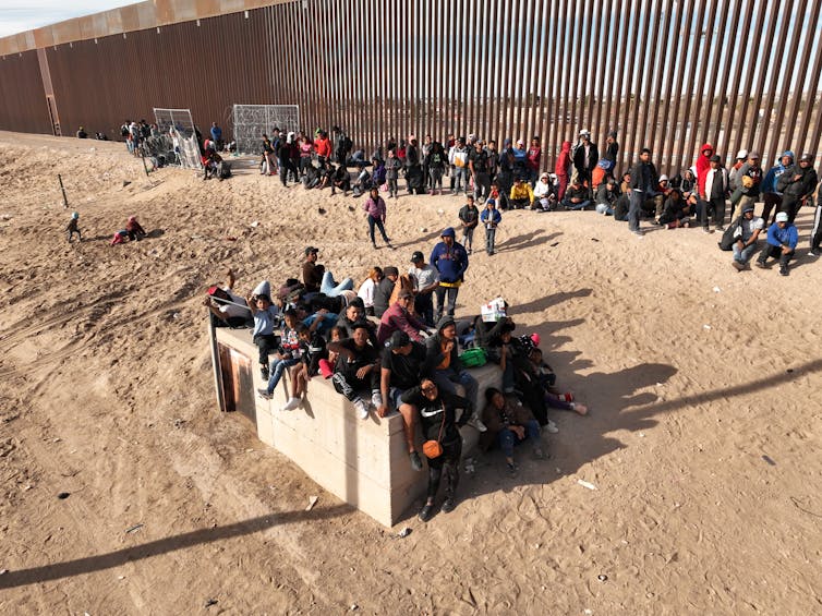 A large group of people are seen sitting and standing along a tall brown fence in an empty area of brown dirt.