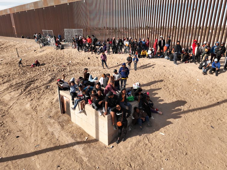 Se ve a un gran grupo de personas sentadas y de pie a lo largo de una alta valla marrón en un área vacía de tierra marrón.