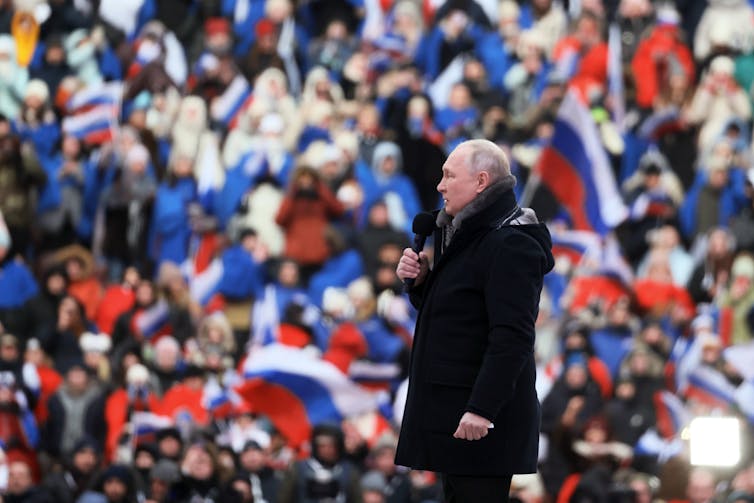 A man holding a microphone stands in front of a crowd waving Russian flags.