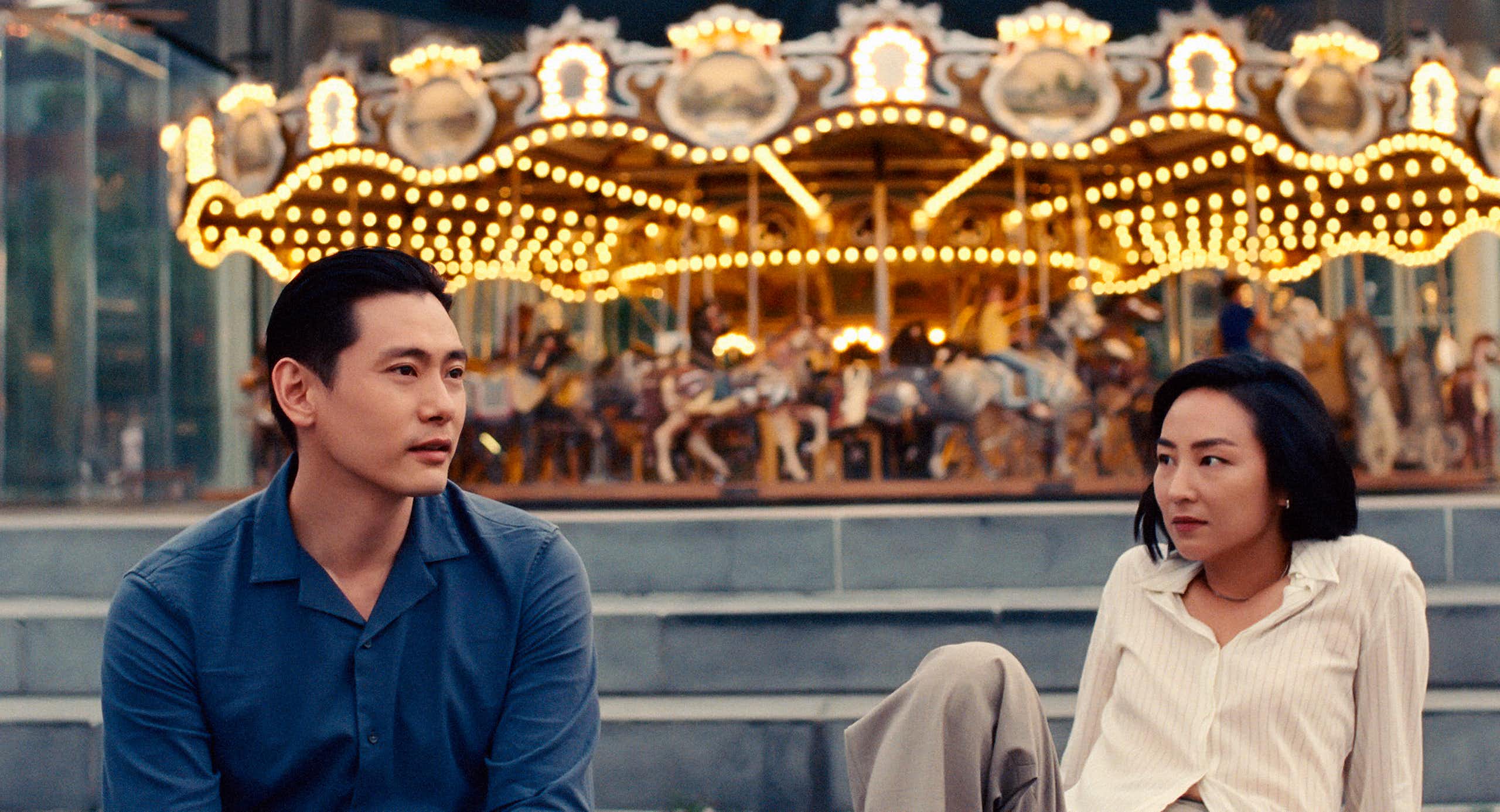 An Asian man and woman sit in front of a merry-go-round