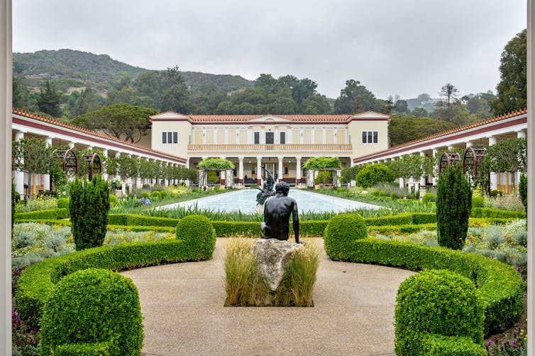 Lush gardens surrounding a Roman villa with a statue seen.