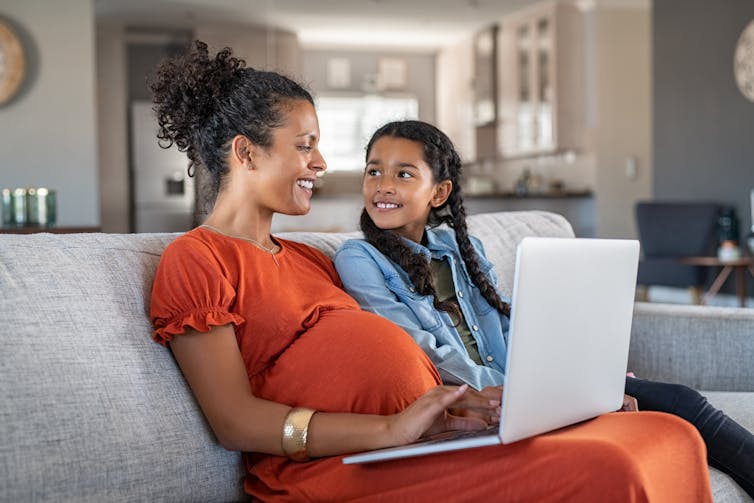 A pregnant woman sits on a couch with her daughter, with a laptop on her lap.