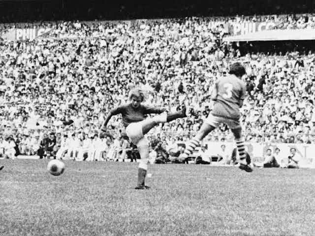 A black and white picture of a woman footballer's shot being blocked in front of a packed stadium.