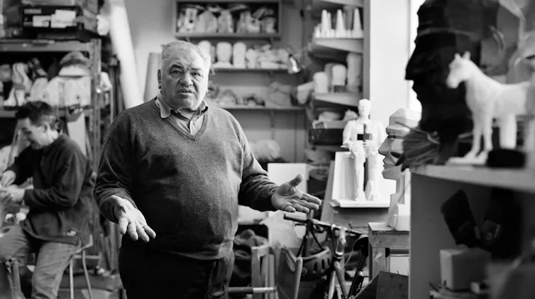 Paolozzi looking at the camera, his arms out and hands upturned, surrounded by sculptures.