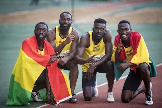 Quatre hommes en tenue d'athlétisme s'agenouillent et s'accroupissent sur une piste de course, deux d'entre eux étant drapés des drapeaux vert, rouge et jaune du Ghana.