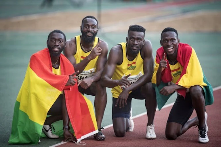 Quatre hommes en tenue d'athlétisme s'agenouillent et s'accroupissent sur une piste de course, deux d'entre eux étant drapés des drapeaux vert, rouge et jaune du Ghana.