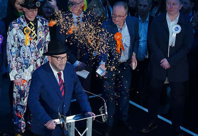 George Galloway giving a speech with other election candidates standing behind him. One is throwing orange confetti over him. 