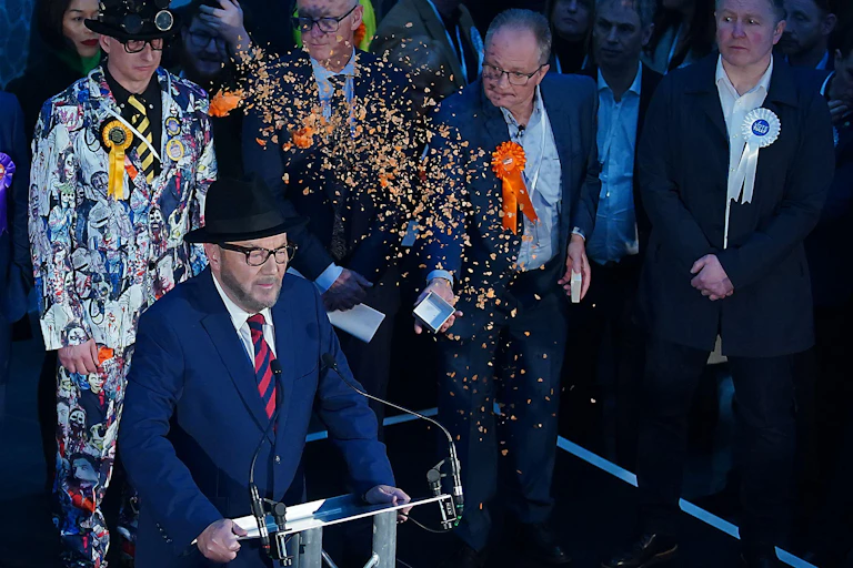 George Galloway giving a speech with other election candidates standing behind him. One is throwing orange confetti over him.