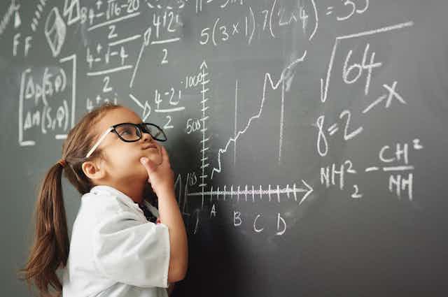 Petite fille avec des lunettes en train de résoudre des équations très compliquées sur un tableau noir dans une classe