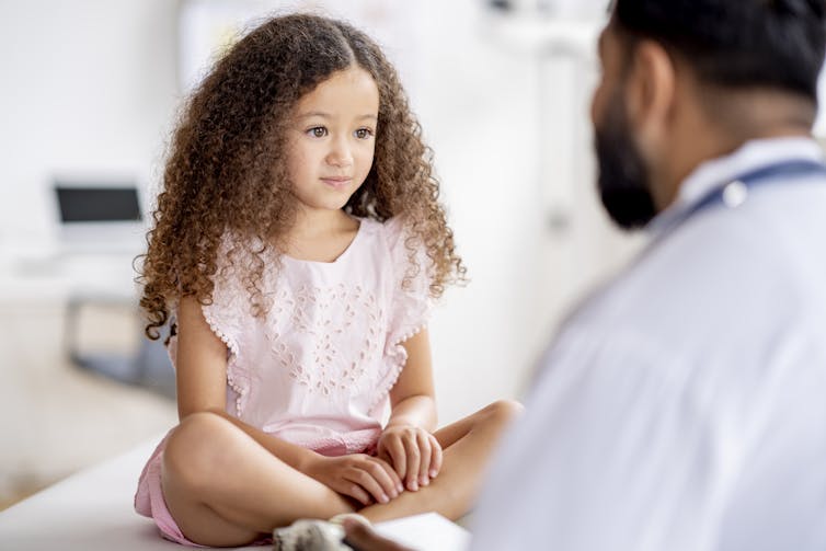 girl sitting on exam table faces a doctor viewer can see from behind