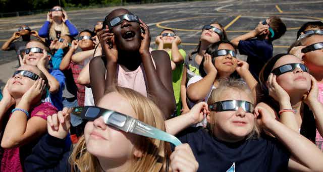 Young students wearing eclipse glasses look up at the sky and smile.