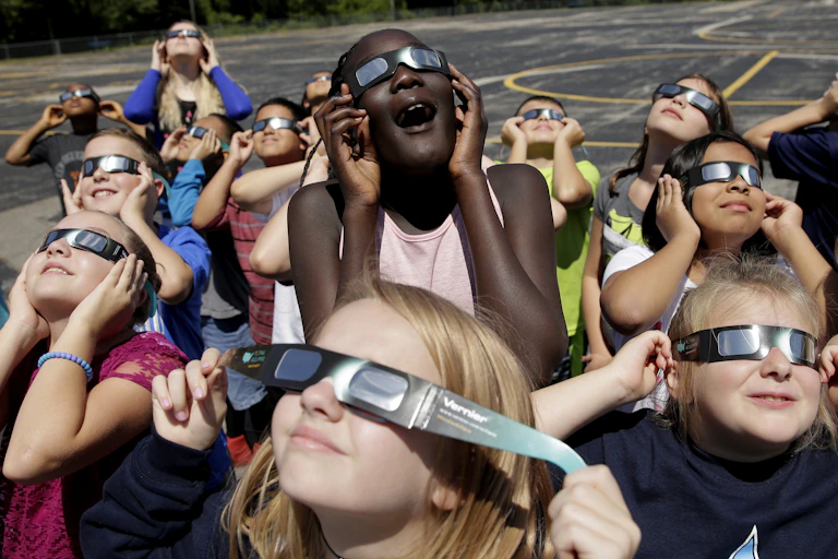Young students wearing eclipse glasses look up at the sky and smile.