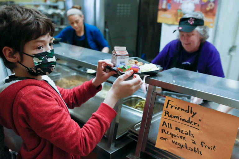 Child wearing mask holding tray of food with two cafeteria workers behind the counter. A sign reads "Friendly reminder: All meals require a fruit or vegetable."