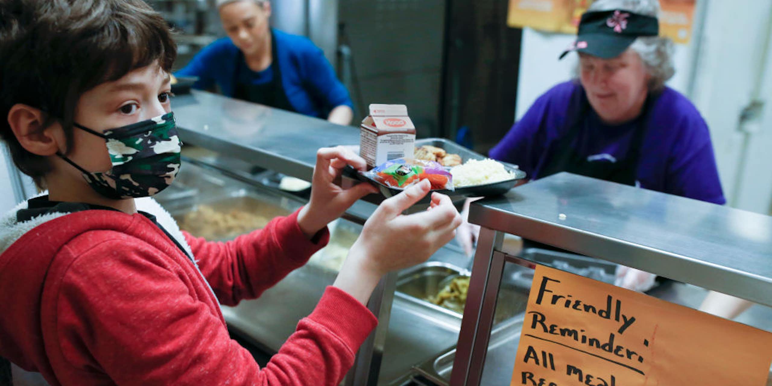 Child wearing mask holding tray of food with two cafeteria workers behind the counter. A sign reads "Friendly reminder: All meals require a fruit or vegetable."