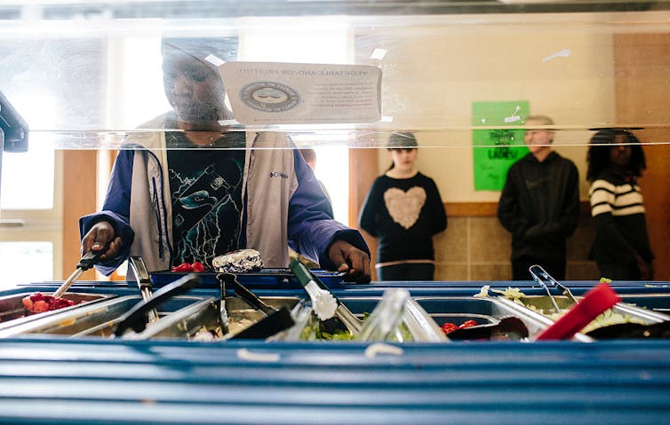 Child scooping food from salad bar onto a tray; other children lean against the wall