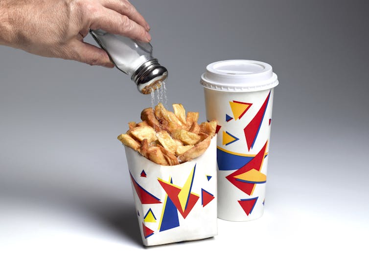 Hand shaking salt on a packet of fries beside a soft drink