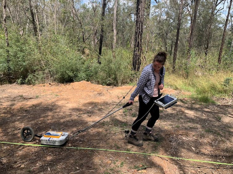 A woman in open bushland dragging an electrical tool across the ground.