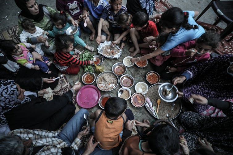 Several children and adults share a meal while being seated in a circle on the floor where a number of dishes are placed in the center.