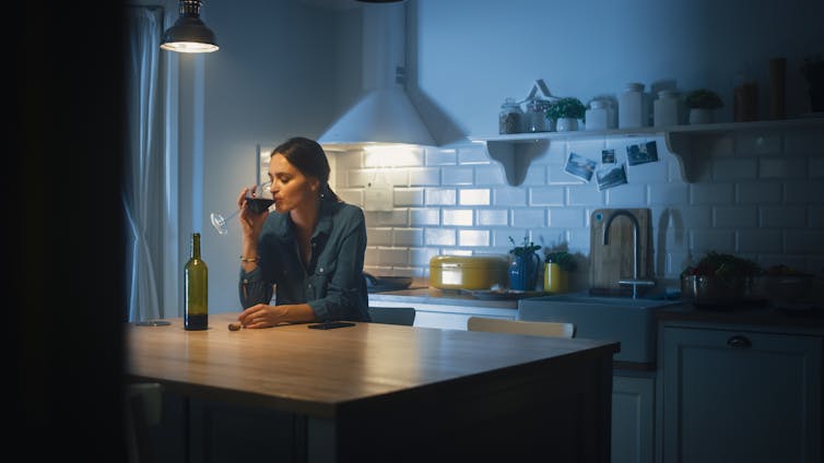 A woman at a kitchen bench drinking a glass of red wine.