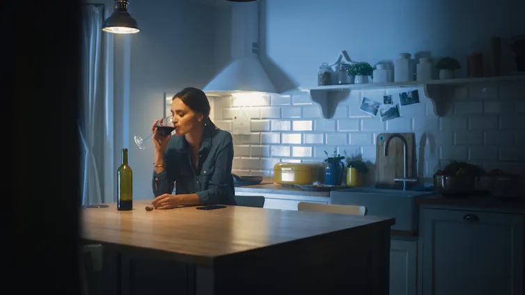A woman at a kitchen bench drinking a glass of red wine.
