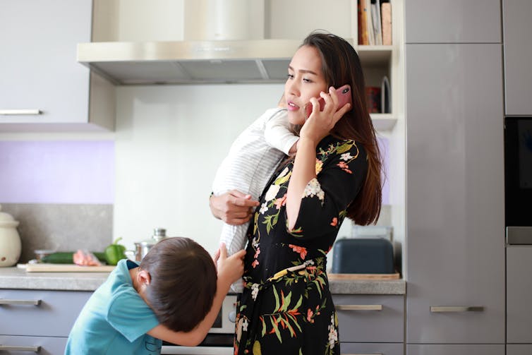 A woman in the kitchen with two children talking on the phone.