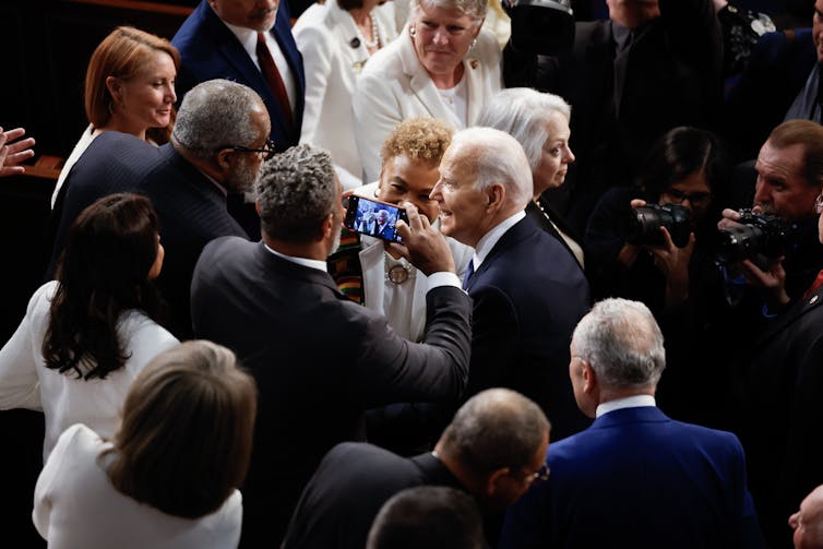 President Joe Biden stands surrounded by people in formal clothing and smiles. One man holds a cell phone camera close up to his face.
