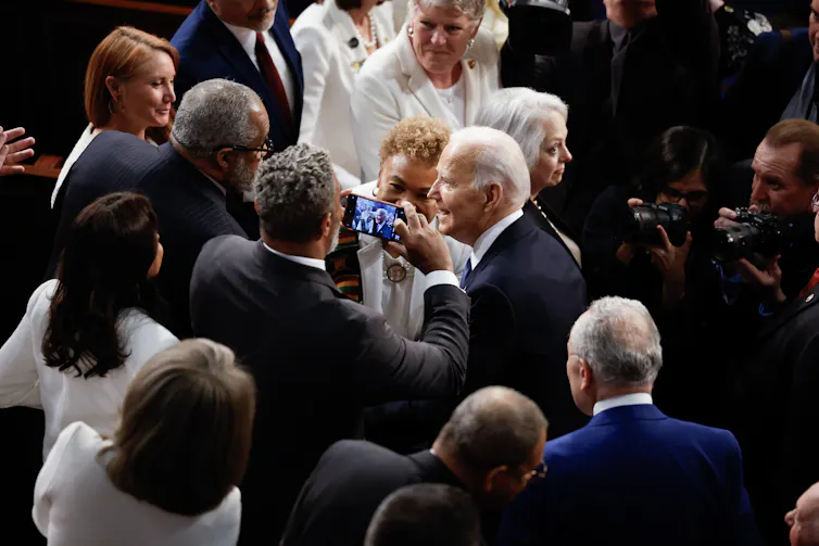President Joe Biden stands surrounded by people in formal clothing and smiles. One man holds a cell phone camera close up to his face.
