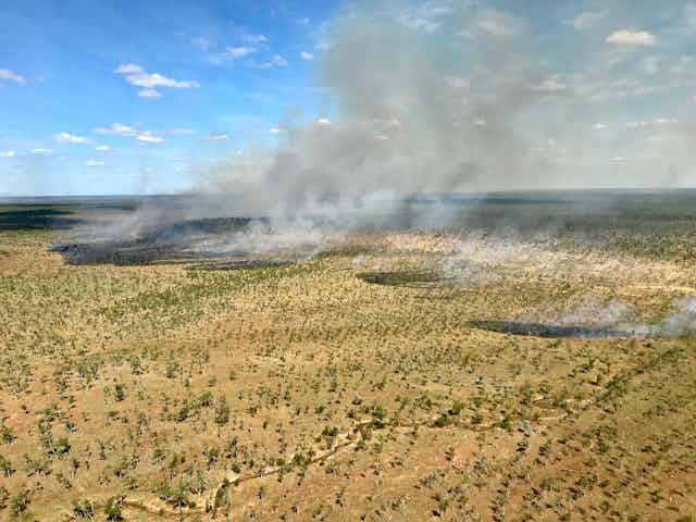 Aerial photo showing small patches of fire and burning with clouds of smoke in a sparsely wooded grassy landscape.