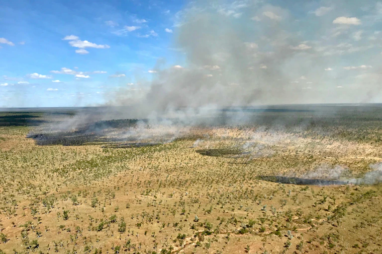 Aerial photo showing small patches of fire and burning with clouds of smoke in a sparsely wooded grassy landscape.