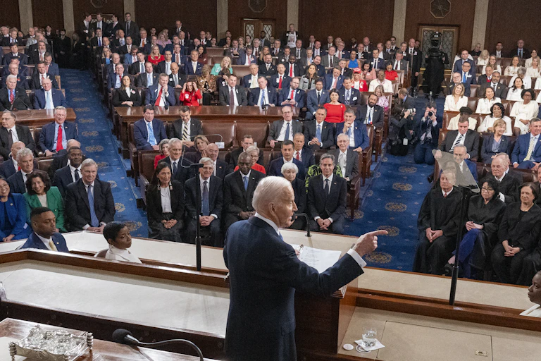 Joe Biden stands at a lectern and points his finger facing a large crowd of people dressed in formal clothing.