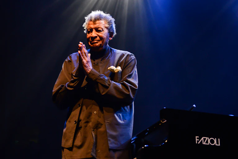 An elderly man with grey hair stands under a light on a stage next to a piano, hands in a thank-you prayer position in front of him.